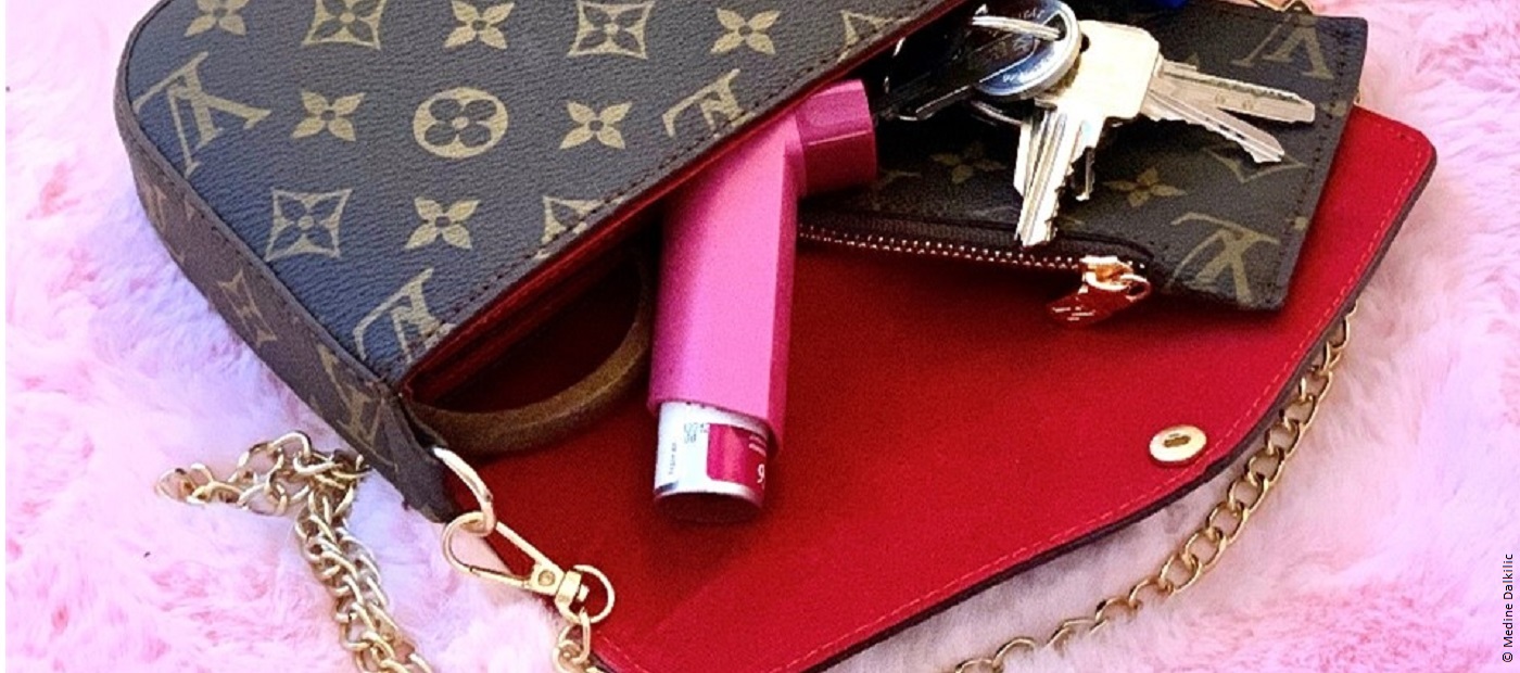 Eine Louis Vuitton Tasche liegt geöffnet auf einem rosa Fell. In der Tasche sind Schlüssel und ein Inhalator zu sehen.  
